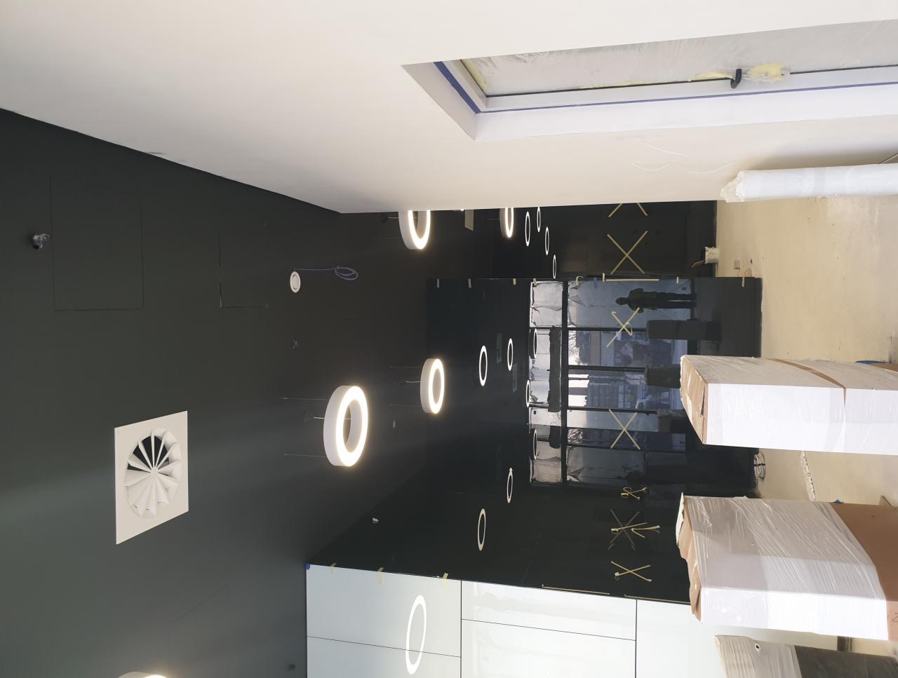 Ciemne ściany biura z okrągłymi lampami LED, biały sufit z wentylacją, zabezpieczone folią okna i materiały budowlane na podłodze.