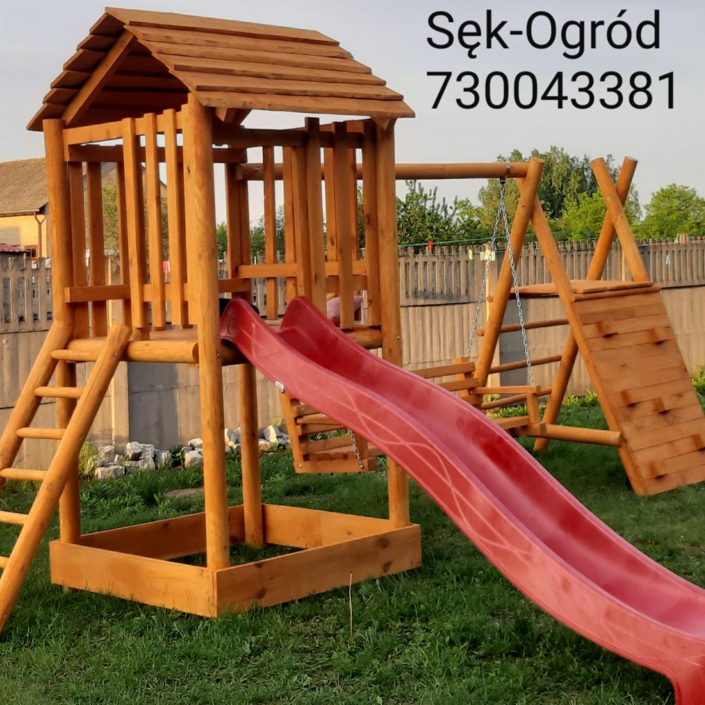 Drewniany plac zabaw dla dzieci z czerwoną zjeżdżalnią, drabinką, huśtawką i ścianką wspinaczkową, widoczny na trawniku przed domem, z numerem telefonu firmy na górze zdjęcia.