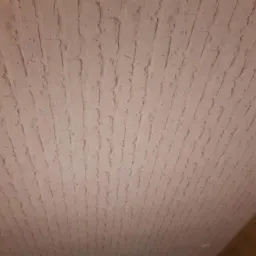 Jasny sufit pokryty świeżo nałożoną, strukturalną gładzią imitującą cegłę, widoczne niedoskonałości i nierówności faktury. Kabel elektryczny i fragment drewnianej podłogi w rogu.