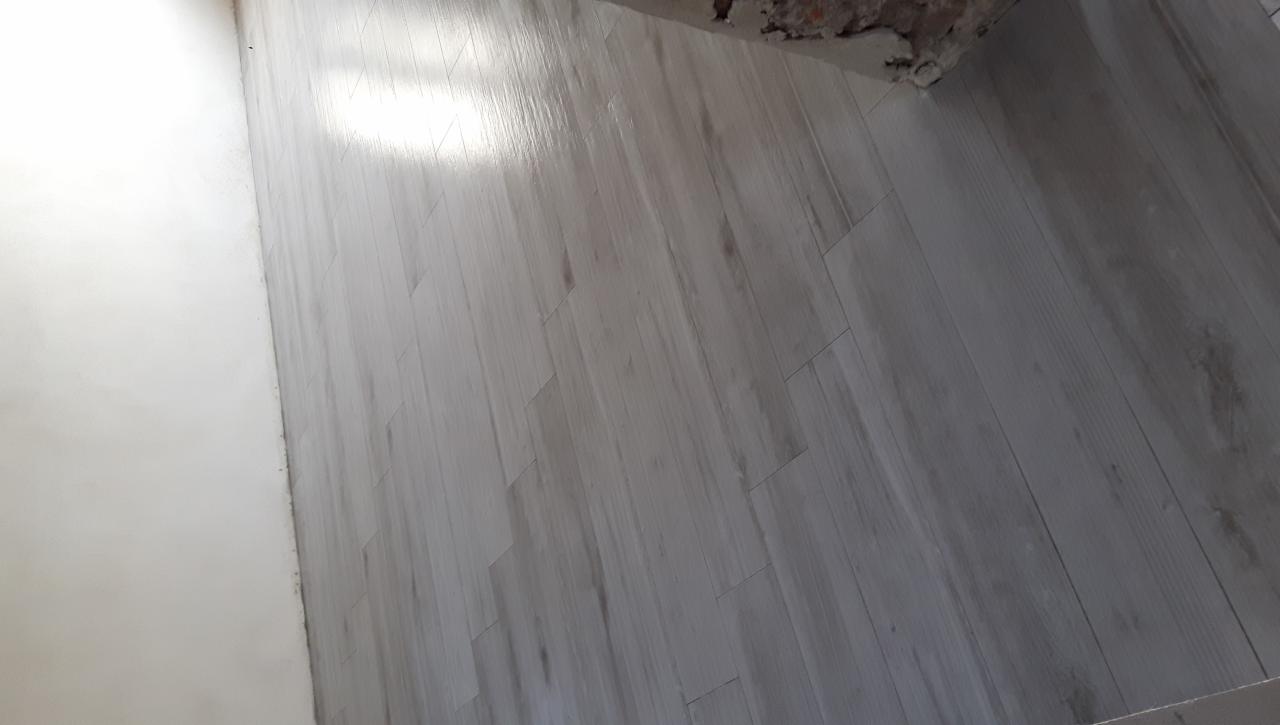 Ułożona podłoga z jasnych, drewnopodobnych płytek, z fragmentem białej ściany po lewej i surowego muru w rogu u góry.