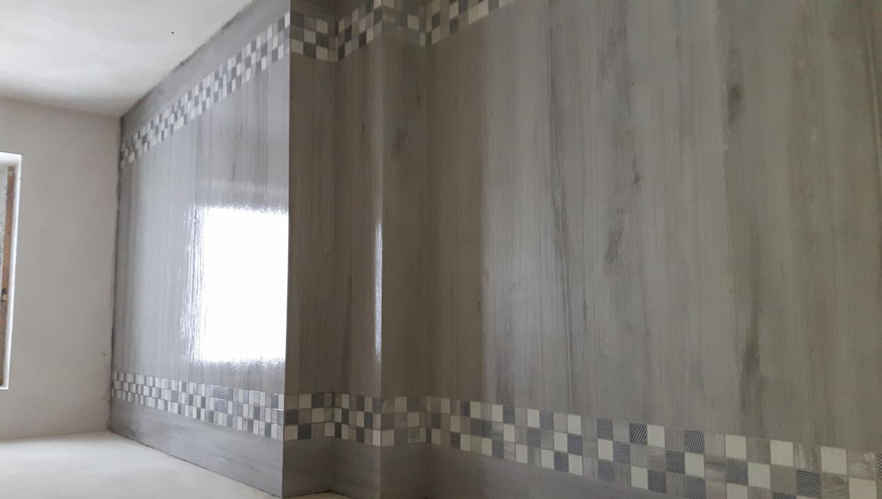 Ściana łazienki wyłożona szarymi płytkami imitującymi drewno z dekoracyjnym pasem mozaiki w szachownicę u góry i na dole, widok z perspektywy narożnika.