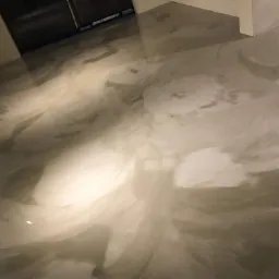 Błyszcząca, szara podłoga z efektem marmuru, wykonana w technologii bezspoinowej, widoczna w rogu pomieszczenia z białymi ścianami i czarnym otworem drzwiowym.