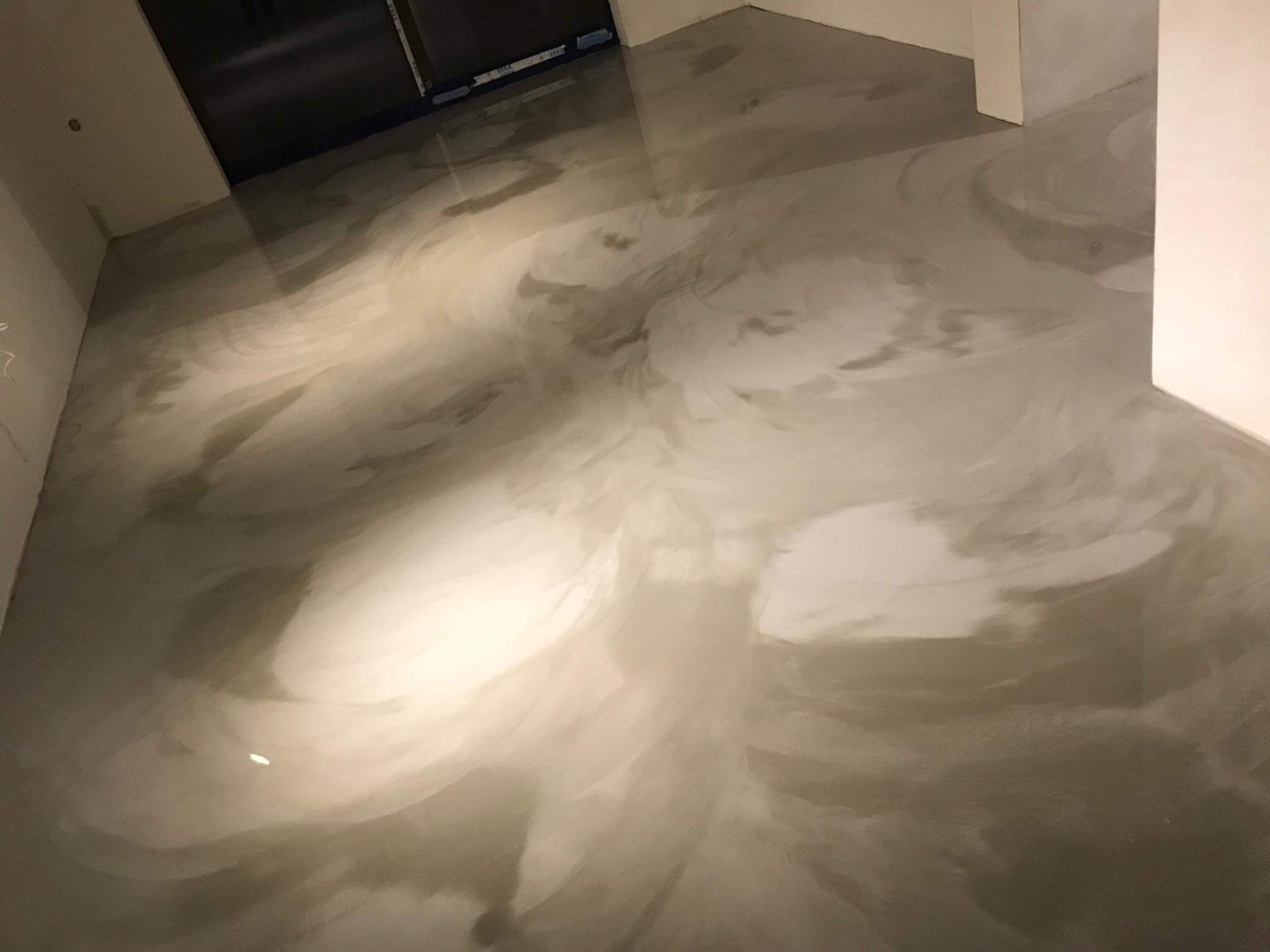 Błyszcząca, szara podłoga z efektem marmuru, wykonana w technologii bezspoinowej, widoczna w rogu pomieszczenia z białymi ścianami i czarnym otworem drzwiowym.