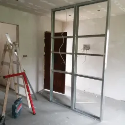 Konstrukcja metalowa z profili, prawdopodobnie pod ściankę działową z płyt gipsowo-kartonowych, widoczna na tle surowych ścian i podłogi w trakcie prac wykończeniowych, drabina i poziomica obok.