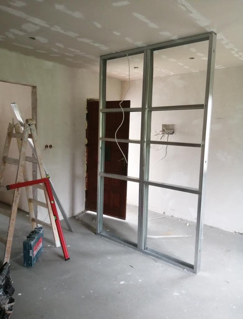 Konstrukcja metalowa z profili, prawdopodobnie pod ściankę działową z płyt gipsowo-kartonowych, widoczna na tle surowych ścian i podłogi w trakcie prac wykończeniowych, drabina i poziomica obok.