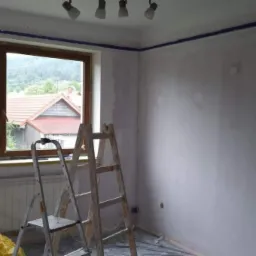 Pokój w trakcie przygotowania do malowania: ściany po wstępnym szpachlowaniu, zabezpieczone taśmą malarską przy suficie, drabiny malarskie ustawione przy oknie z widokiem na zieleń.