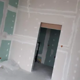 Wnętrze w trakcie montażu płyt karton-gips, widoczne zielone płyty częściowo pokryte szpachlą, wystające kable elektryczne i otwór drzwiowy z widoczną konstrukcją.