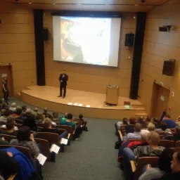 Prelegent w garniturze prezentuje na scenie przed publicznością w sali konferencyjnej z projektorem wyświetlającym obraz.