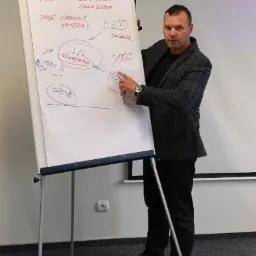 Mężczyzna w szarej marynarce prezentuje wykres na flipcharcie z napisem 'PREMA KOSCIOWA JAKOSCIOWA' i zaznaczonymi wartościami 700% i 3000.