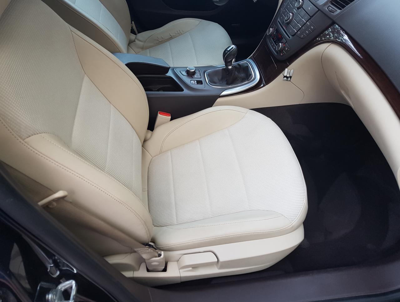 Jasna, beżowa tapicerka fotela samochodowego z widocznym fragmentem deski rozdzielczej i dźwignią zmiany biegów. Fotel z wyraźną fakturą materiału i przeszyciami.