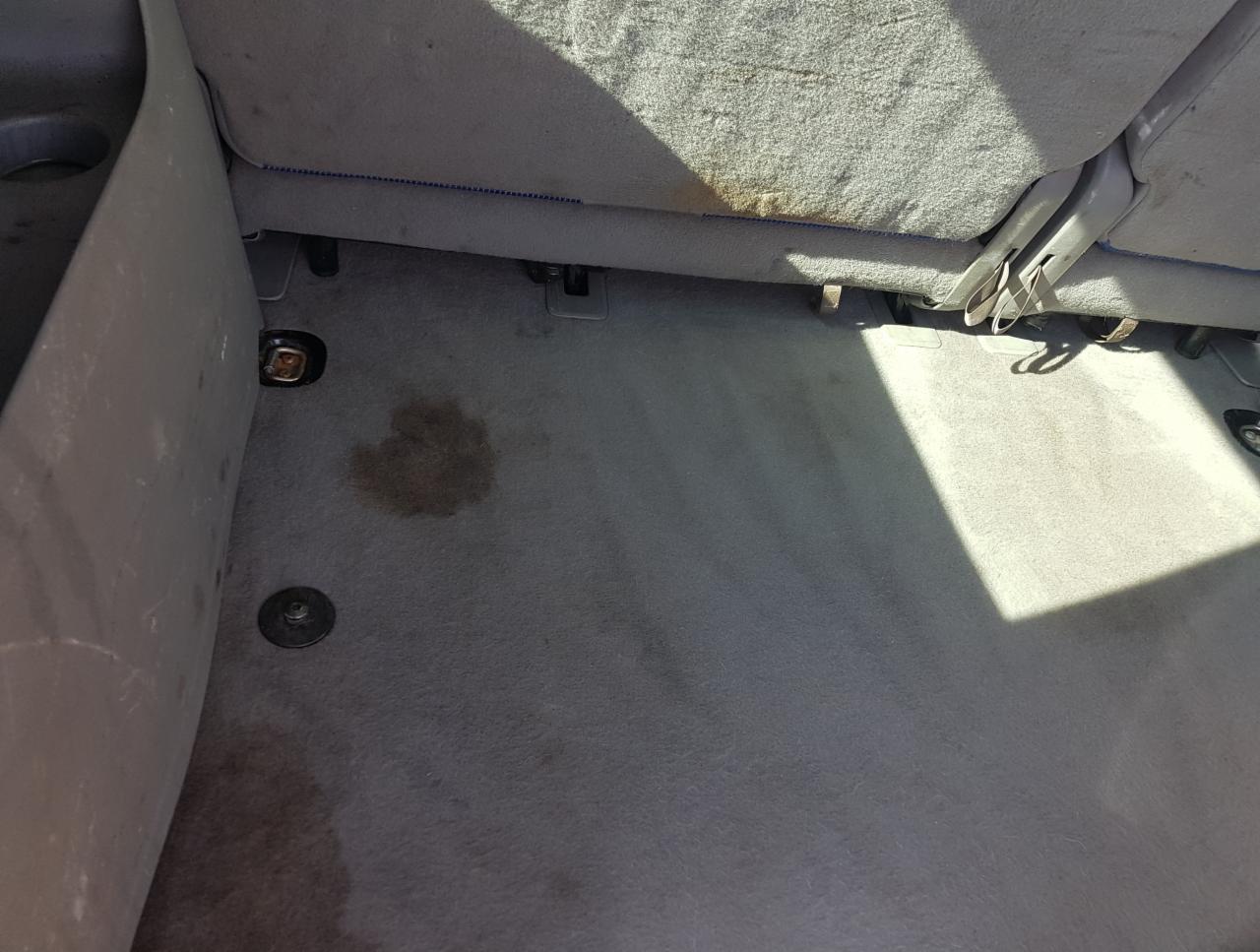 Brudna, szara tapicerka samochodowa z plamami i zaciekami, widoczne mocowania siedzeń i fragment oparcia.