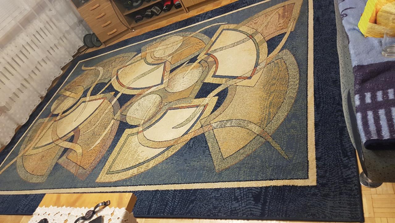 Duży, prostokątny dywan z abstrakcyjnym wzorem w odcieniach beżu, brązu i niebieskiego, leżący na drewnianej podłodze. W tle widoczna szafka na buty i zasłona.