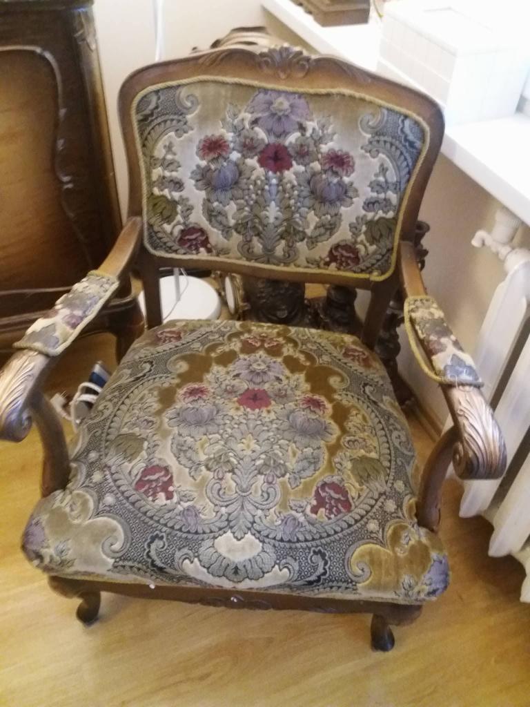 Antyczny fotel z bogato zdobioną tapicerką w kwiaty i ornamenty, widoczny na tle drewnianej komody i grzejnika.