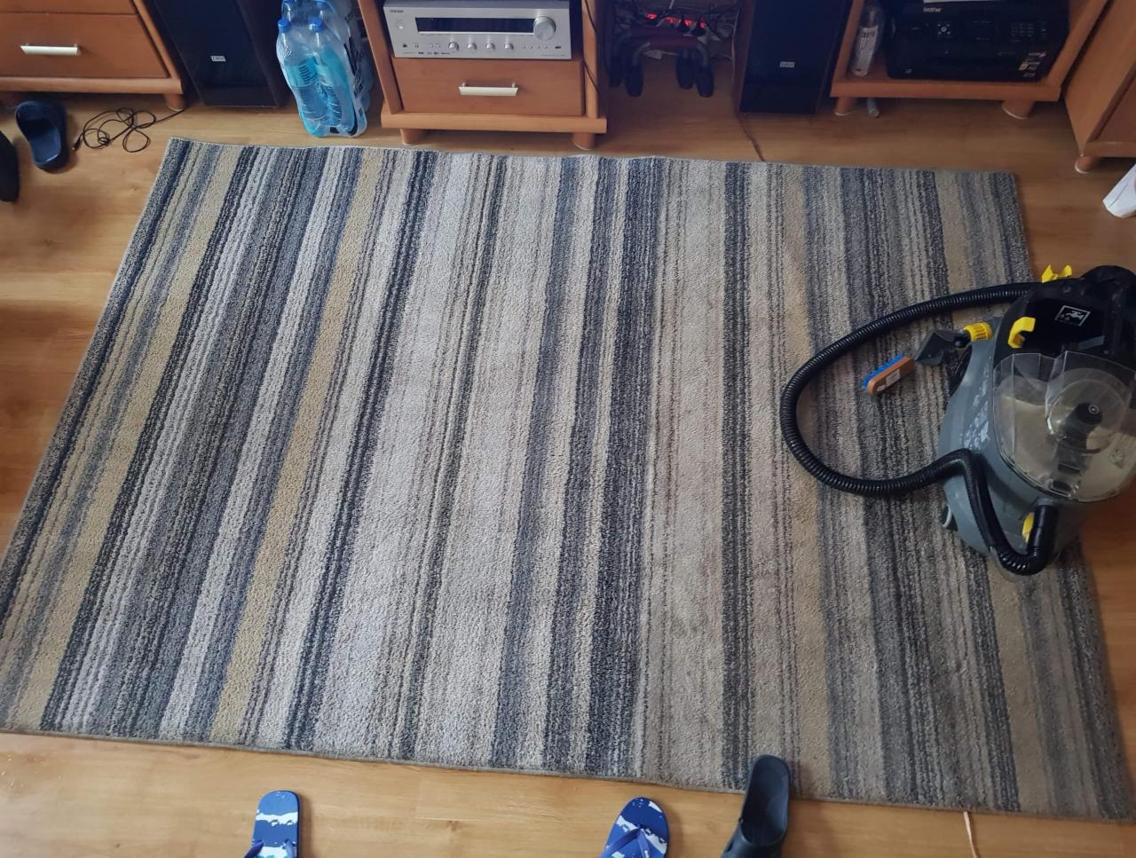 Pasiasty dywan w odcieniach szarości i beżu, częściowo wyczyszczony, z widocznym odkurzaczem piorącym i fragmentami obuwia na podłodze.