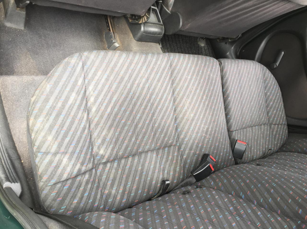 Tylna kanapa samochodu z szaro-czarną tapicerką w paski, z widocznymi pasami bezpieczeństwa, lekko zakurzona.