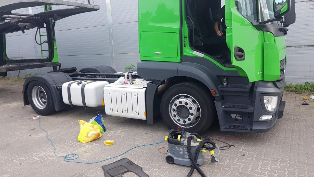 Zielony ciągnik siodłowy z odpiętą naczepą, widoczny od boku, obok stoi odkurzacz piorący i leżą wyprane dywaniki samochodowe, na zbiorniku paliwa stoją środki czyszczące.