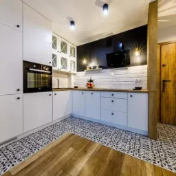 Nowoczesna kuchnia z białymi szafkami, czarnym akcentem na ścianie, białymi płytkami metra, wzorzystą podłogą i drewnianymi elementami, oświetlona trzema lampami sufitowymi.