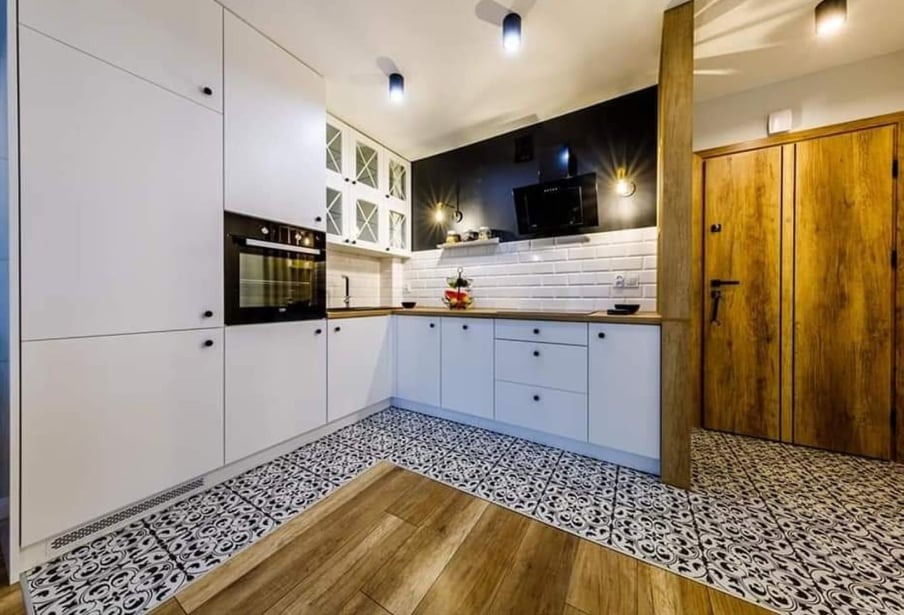 Nowoczesna kuchnia z białymi szafkami, czarnym akcentem na ścianie, białymi płytkami metra, wzorzystą podłogą i drewnianymi elementami, oświetlona trzema lampami sufitowymi.