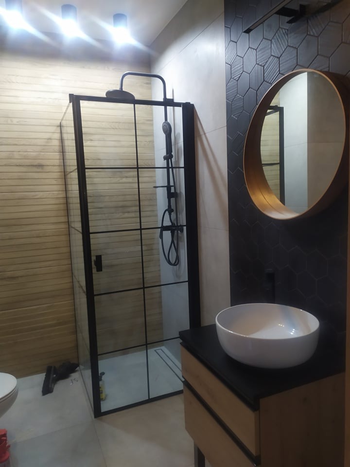 Nowoczesna łazienka z kabiną prysznicową typu walk-in z czarnymi profilami, umywalką nablatową i lustrem w okrągłej ramie. Ściana wykończona płytkami heksagonalnymi.