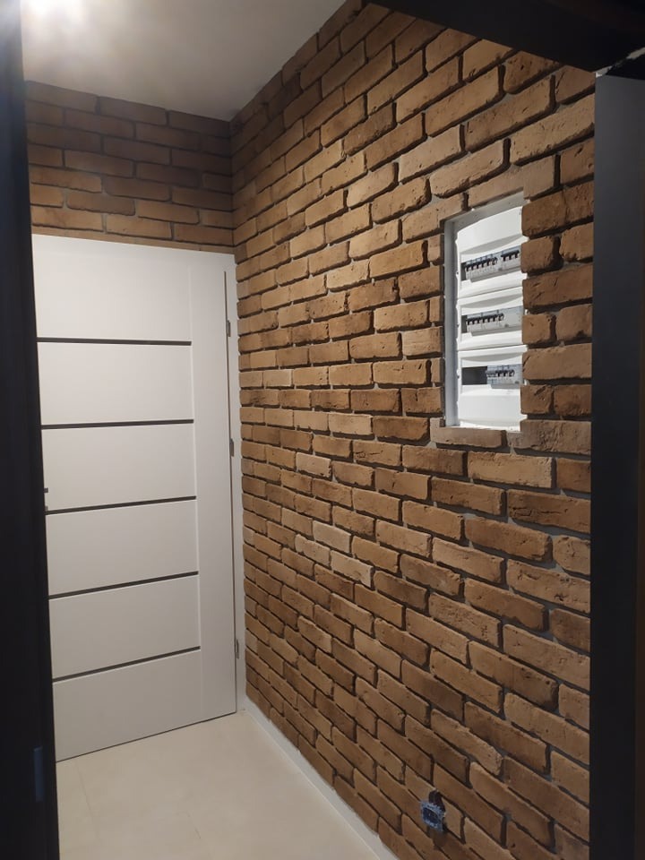 Ściana z cegły dekoracyjnej w odcieniach brązu, z widoczną białą skrzynką elektryczną i fragmentem białych drzwi z czarnymi paskami, całość w nowoczesnym wnętrzu.