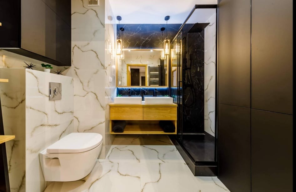 Nowoczesna łazienka z białymi płytkami imitującymi marmur, drewnianą szafką pod umywalką z dwoma białymi umywalkami, czarną kabiną prysznicową typu walk-in i podwieszaną toaletą.