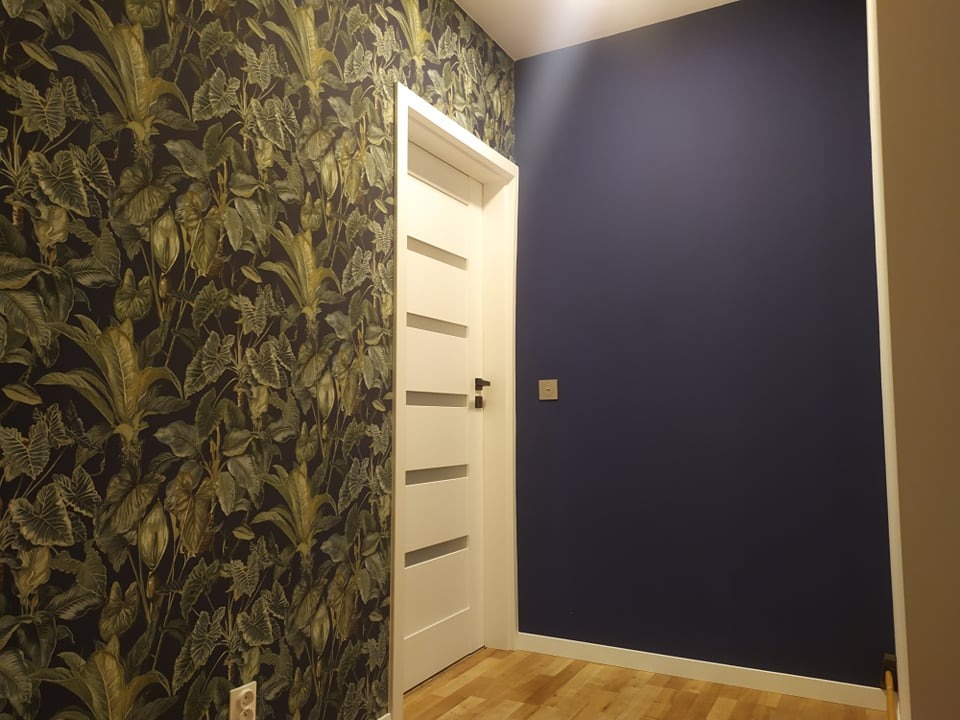 Wnętrze mieszkania po remoncie: ściana pokryta tapetą z motywem tropikalnych liści, sąsiadująca z białą ścianą pomalowaną na jednolity, ciemnoniebieski kolor. Widoczna biała, nowoczesna stolarka...