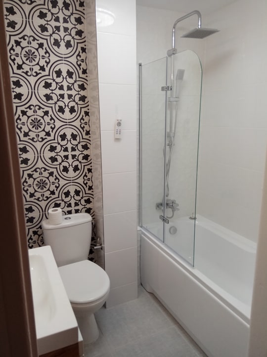 Kompaktowa łazienka z białą armaturą, kabiną prysznicową nad wanną oraz ścianą akcentującą z czarno-białymi płytkami w stylu retro, papier toaletowy na spłuczce.