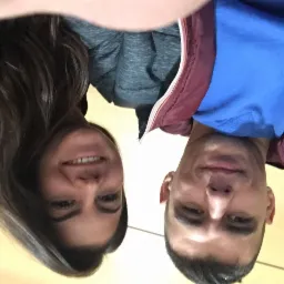 Portret selfie uśmiechniętej kobiety z długimi brązowymi włosami i mężczyzny w niebieskiej bluzie i bordowej kurtce na jasnym tle.