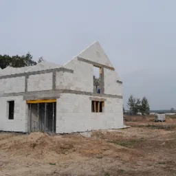 Surowy stan domu w trakcie budowy z betonu kom&oacute;rkowego, widoczne otwory okienne i drzwiowe, zbrojenia nadproży oraz rusztowanie podtrzymujące strop.