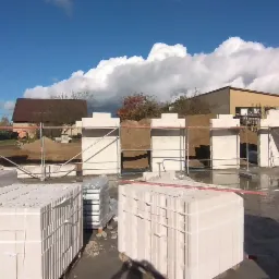 Budowa domu z białych bloczków silikatowych, widoczne rusztowania, pracownicy na budowie, palety z materiałami, w tle domy i błękitne niebo z chmurami.