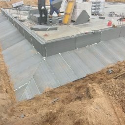 PRBNI ŁK - Budowa fundamentów domu: widok na wykop z izolacją termiczną ścian fundamentowych, pracownik z niebieskim wężem wentylacyjnym, palety z bloczkami silikatowymi w tle.