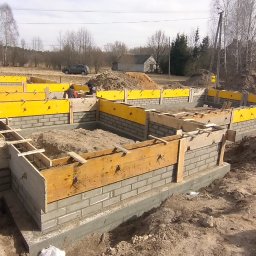 PRBNI ŁK - Wykopane fundamenty z częściowo wzniesionymi ścianami z bloczków betonowych, wzmocnione żółtym szalunkiem i drewnianymi stemplami, widoczne prace murarskie w tle, krajobraz wiejski.