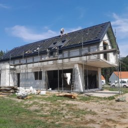 PRBNI ŁK - Budowa domu z białych bloczków: widok z zewnątrz na rusztowania, częściowo położony dach z czarną dachówką i pracującego na nim dekarza, w tle inne budynki i drzewa.