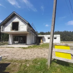 Surowy stan domu z betonu komórkowego w trakcie budowy, widoczne niezadaszone ściany, rusztowania i teren budowy z elementami ogrodzenia z żółtymi tablicami ostrzegawczymi.