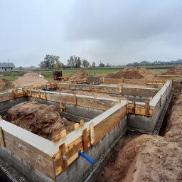PRBNI ŁK - Wylane fundamenty pod budynek jednorodzinny, widoczne szalunki drewniane, ziemia wokół wykopu, na horyzoncie zabudowania.