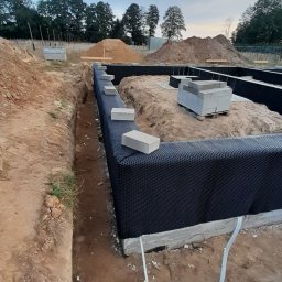 PRBNI ŁK - Fundamenty domu jednorodzinnego zabezpieczone folią kubełkową, widoczne wykopane rowy, przygotowane bloczki betonowe i pryzmy ziemi w tle.