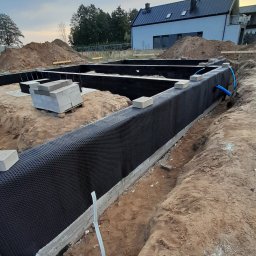 PRBNI ŁK - Wykop pod fundamenty z izolacją pionową z folii kubełkowej, widoczne bloczki betonowe i rury instalacyjne, w tle dom w trakcie budowy.