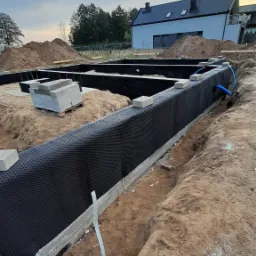 Wykop pod fundamenty z izolacją pionową z folii kubełkowej, widoczne bloczki betonowe i rury instalacyjne, w tle dom w trakcie budowy.