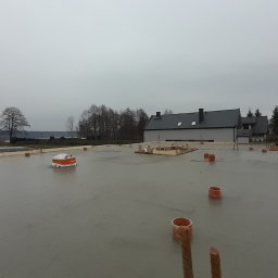 PRBNI ŁK - Wylany fundament pod dom jednorodzinny z widocznymi prętami zbrojeniowymi, deskowaniem i elementami instalacji, na tle zabudowy jednorodzinnej i pochmurnego nieba.