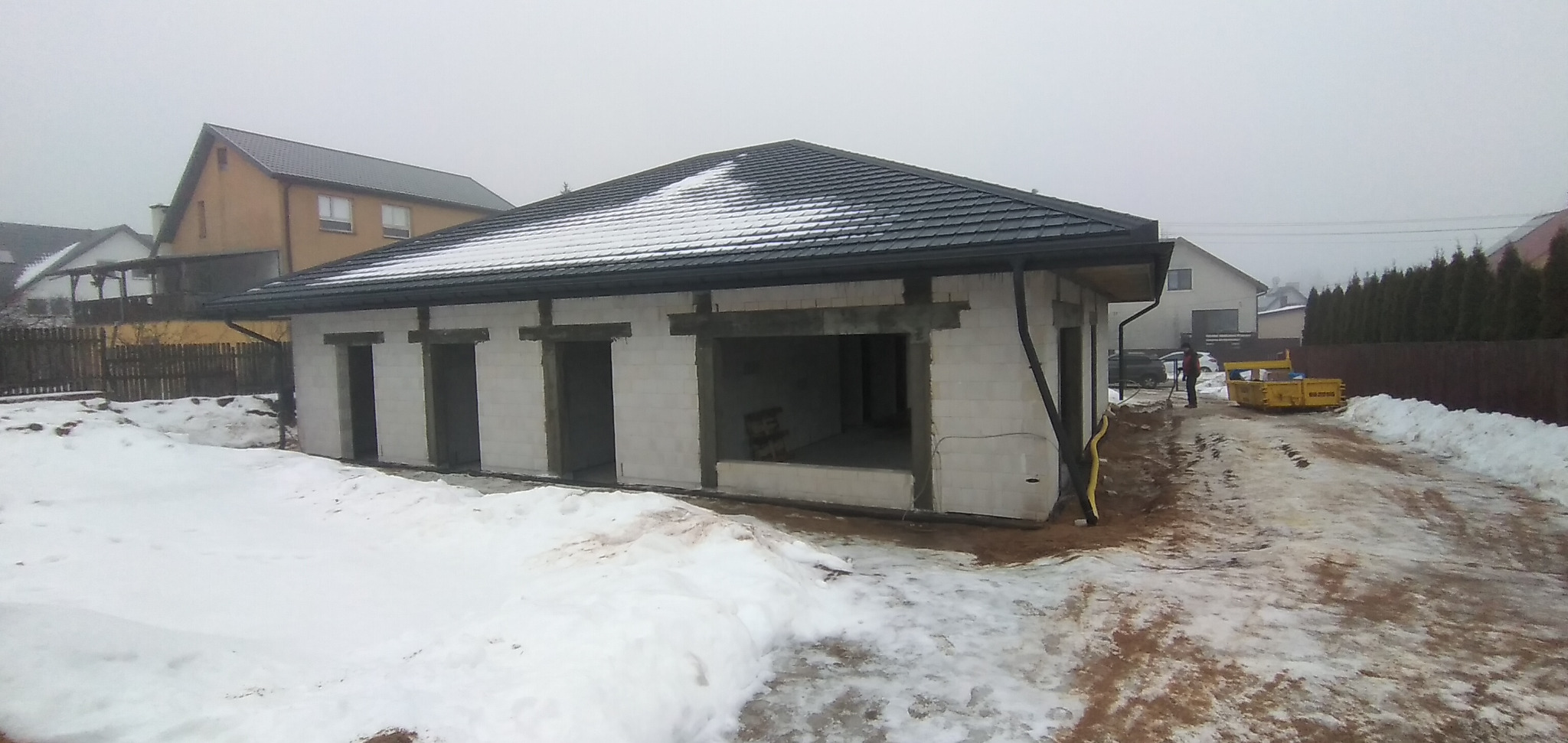 Surowy budynek z białych bloczków, z czarnym dachem pokrytym śniegiem, widoczny żółty wąż i kontener na gruz na zewnątrz, w zimowym krajobrazie.
