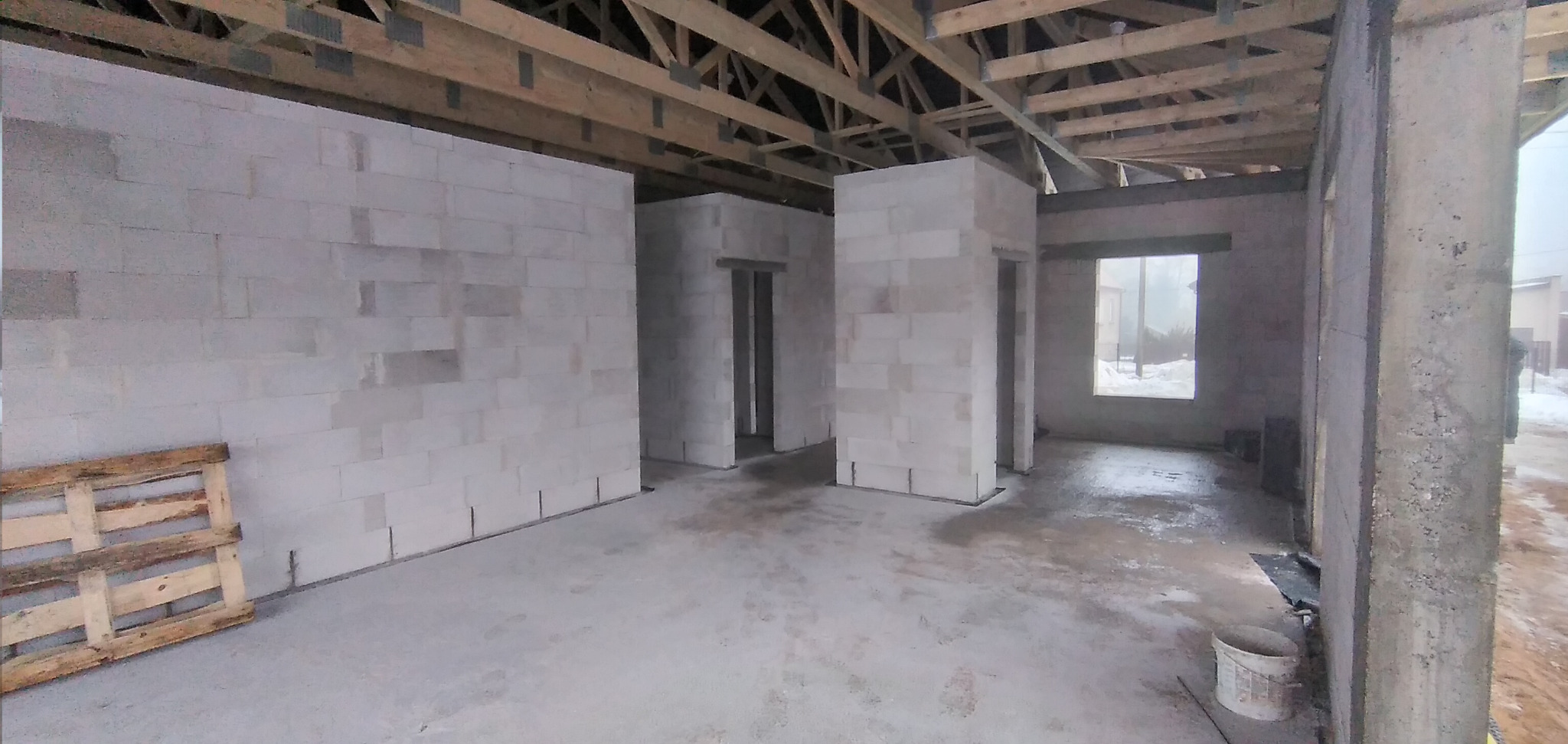 Wnętrze surowego domu w trakcie budowy: ściany z betonu komórkowego, drewniana konstrukcja dachu, widok na zewnątrz przez okno, paleta i wiadro na podłodze.