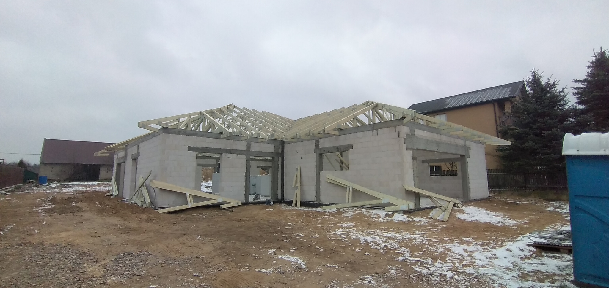 Surowy stan domu z betonu komórkowego z widoczną konstrukcją dachu z drewna, na niezagospodarowanej działce z resztkami śniegu.