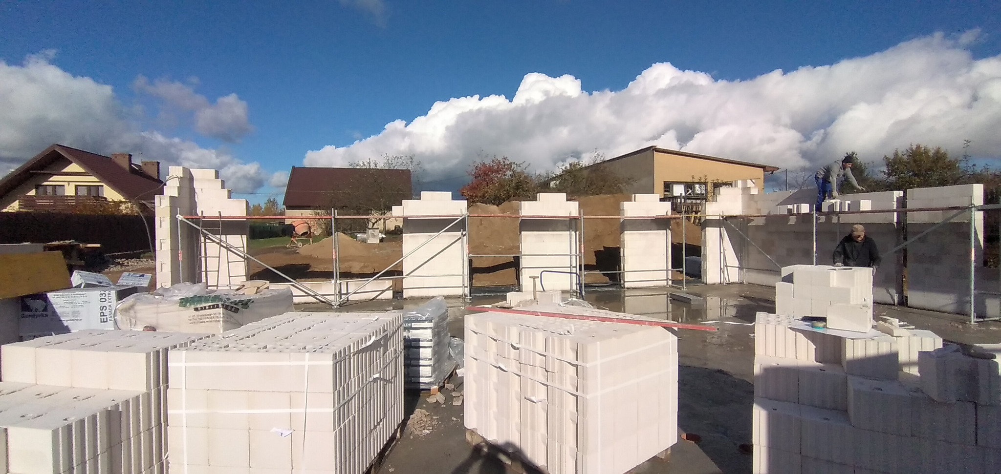 Budowa domu z białych bloczków silikatowych, widoczne rusztowania, pracownicy na budowie, palety z materiałami, w tle domy i błękitne niebo z chmurami.