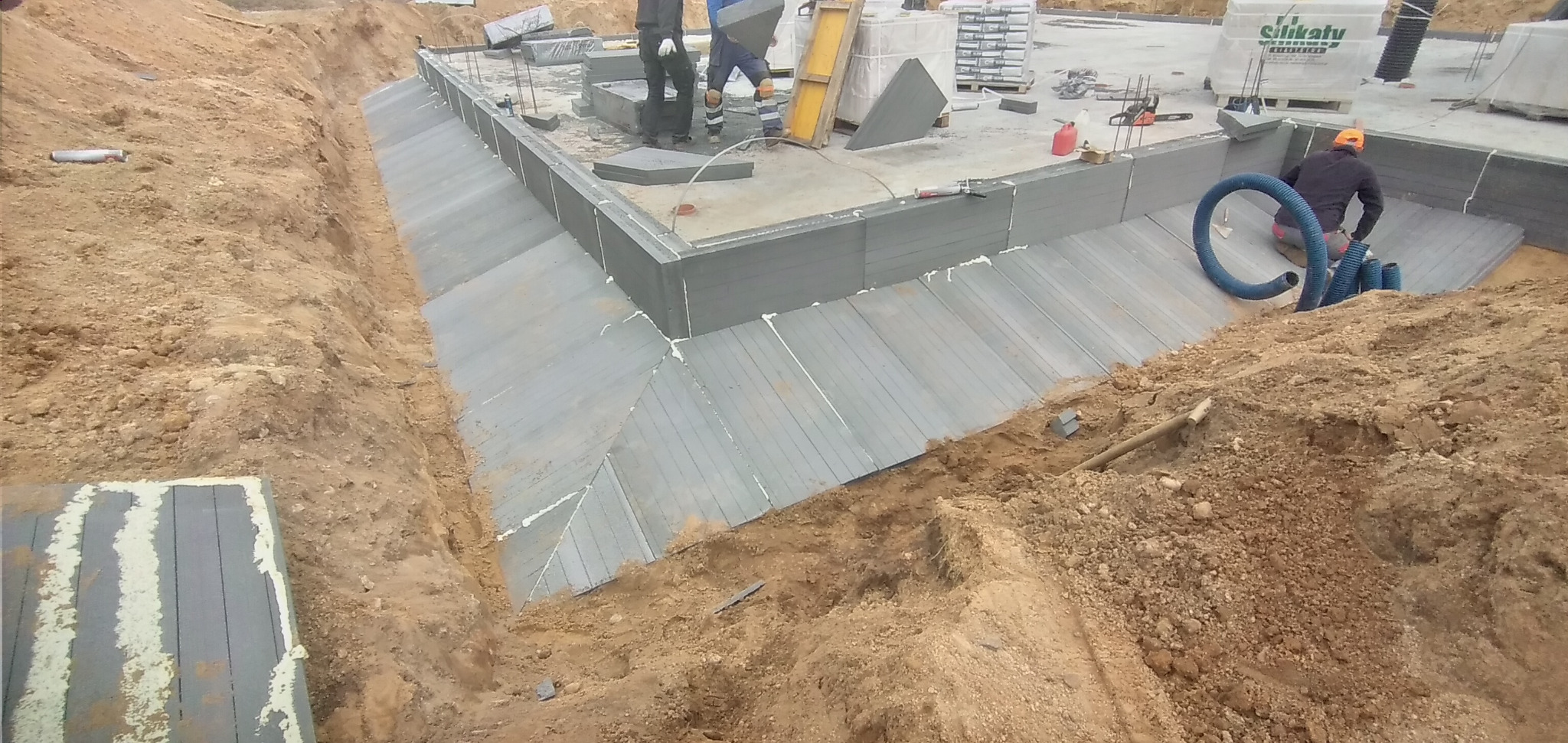 Budowa fundamentów domu: widok na wykop z izolacją termiczną ścian fundamentowych, pracownik z niebieskim wężem wentylacyjnym, palety z bloczkami silikatowymi w tle.