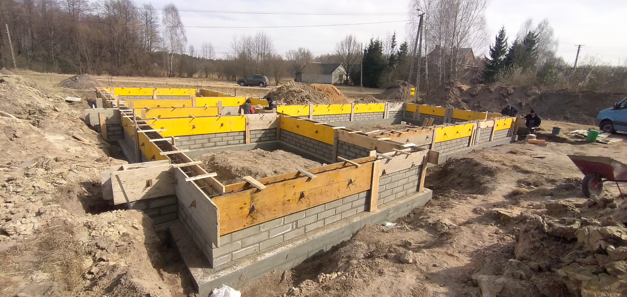 Wykopane fundamenty z częściowo wzniesionymi ścianami z bloczków betonowych, wzmocnione żółtym szalunkiem i drewnianymi stemplami, widoczne prace murarskie w tle, krajobraz wiejski.