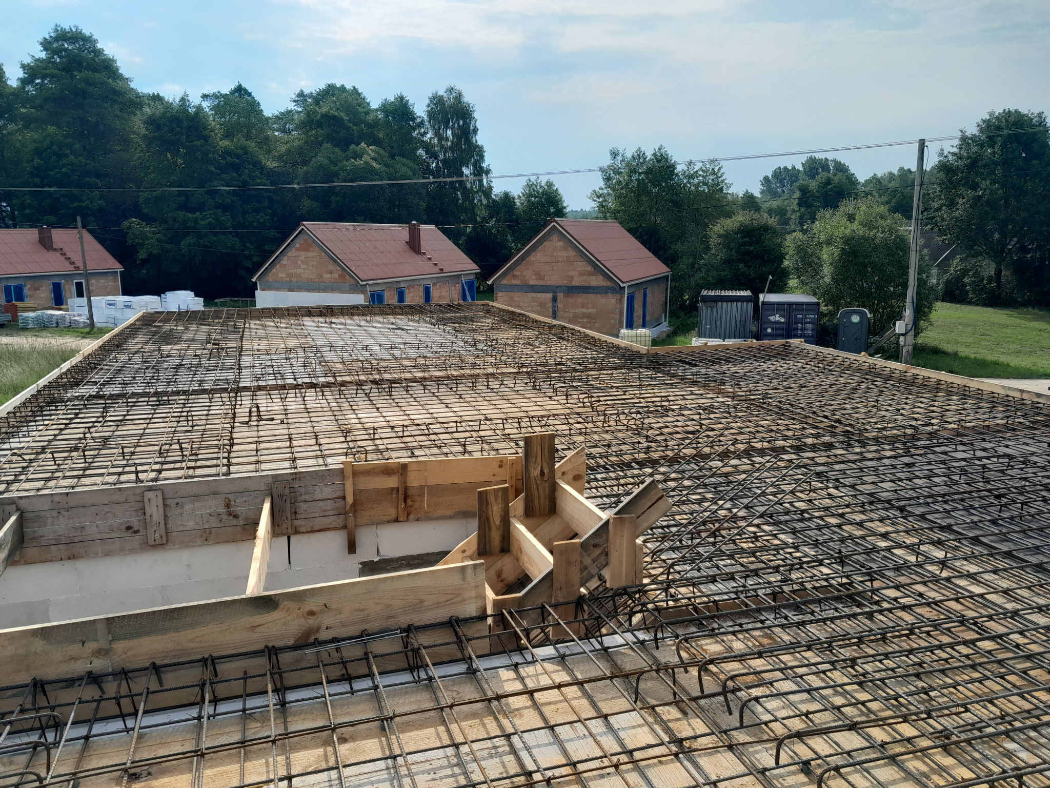 Siatka zbrojeniowa na fundamencie płyty, drewniane szalunki, w tle budynki w stanie surowym otwarte, częściowo pokryte czerwoną dachówką.