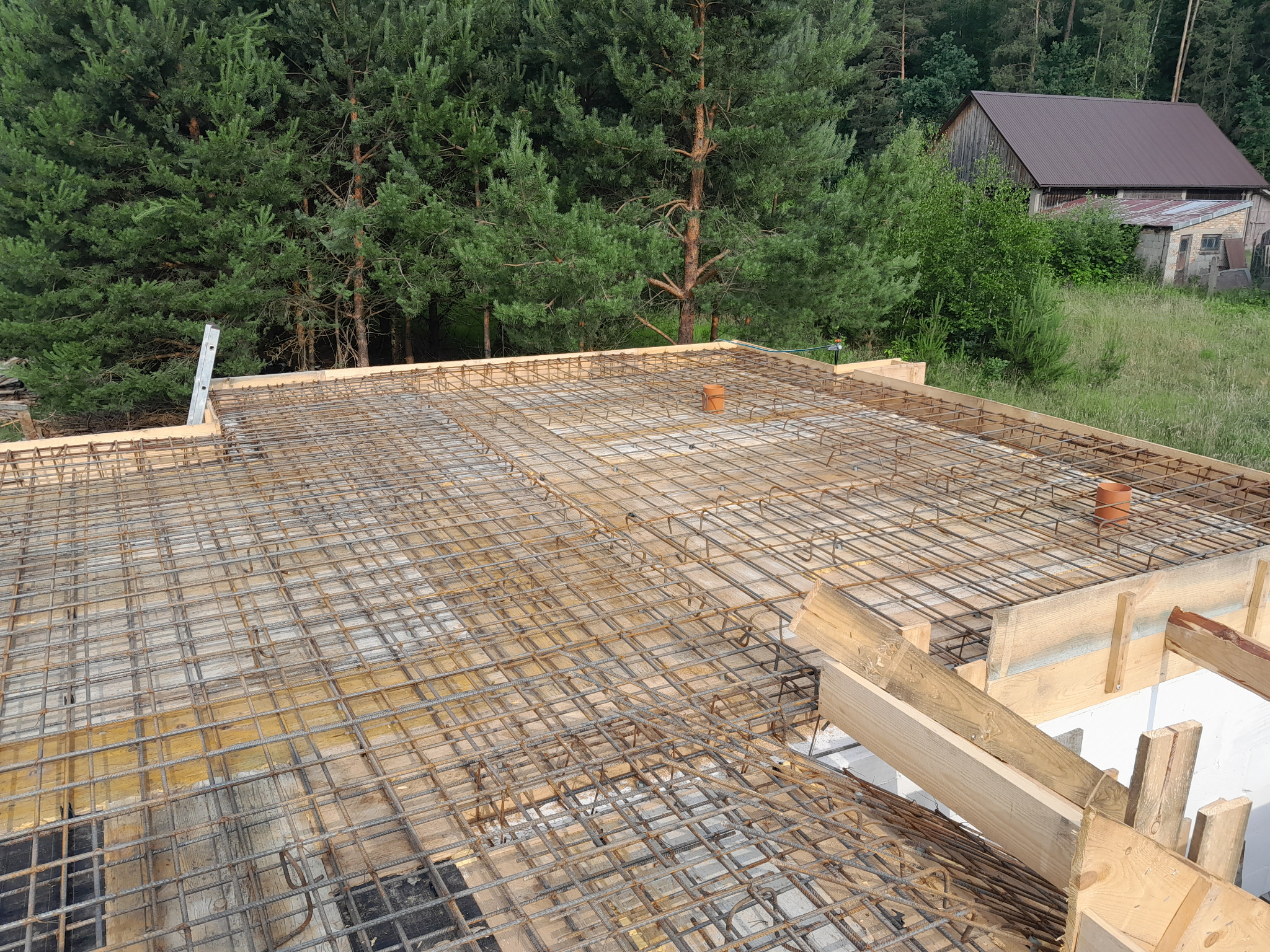 Zbrojenie płyty fundamentowej z drewnianym szalunkiem, widok z góry na tle lasu i zabudowań gospodarczych.