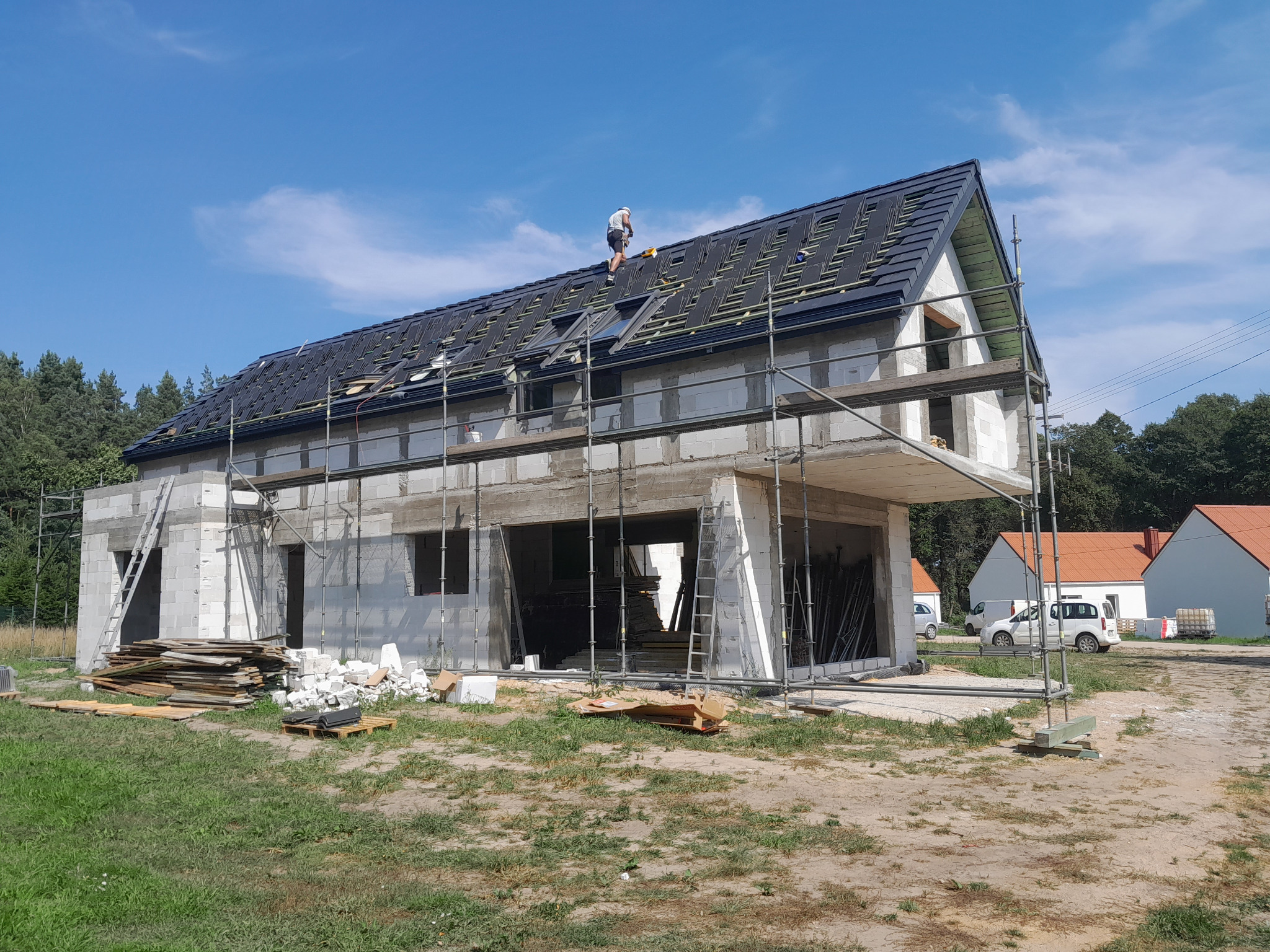 Budowa domu z białych bloczków: widok z zewnątrz na rusztowania, częściowo położony dach z czarną dachówką i pracującego na nim dekarza, w tle inne budynki i drzewa.
