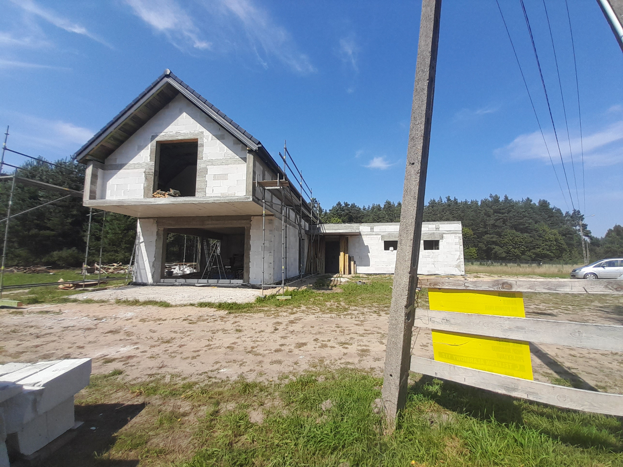 Surowy stan domu z betonu komórkowego w trakcie budowy, widoczne niezadaszone ściany, rusztowania i teren budowy z elementami ogrodzenia z żółtymi tablicami ostrzegawczymi.