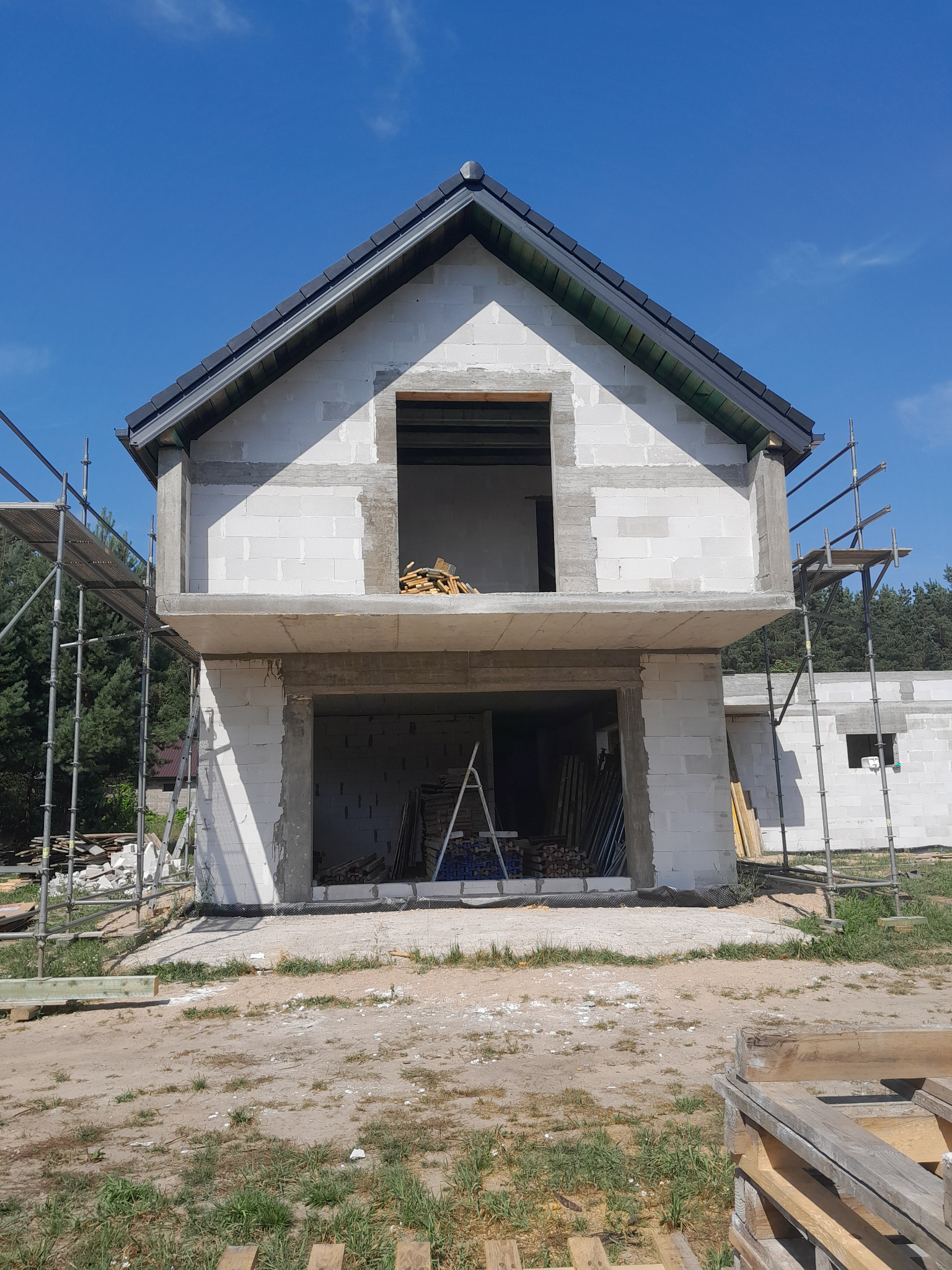 Surowy stan dwukondygnacyjnego domu z białych bloczków, z widocznym zbrojonym stropem i częściowo wykonanym dachem z czarnej dachówki, otoczony rusztowaniami i placem budowy.